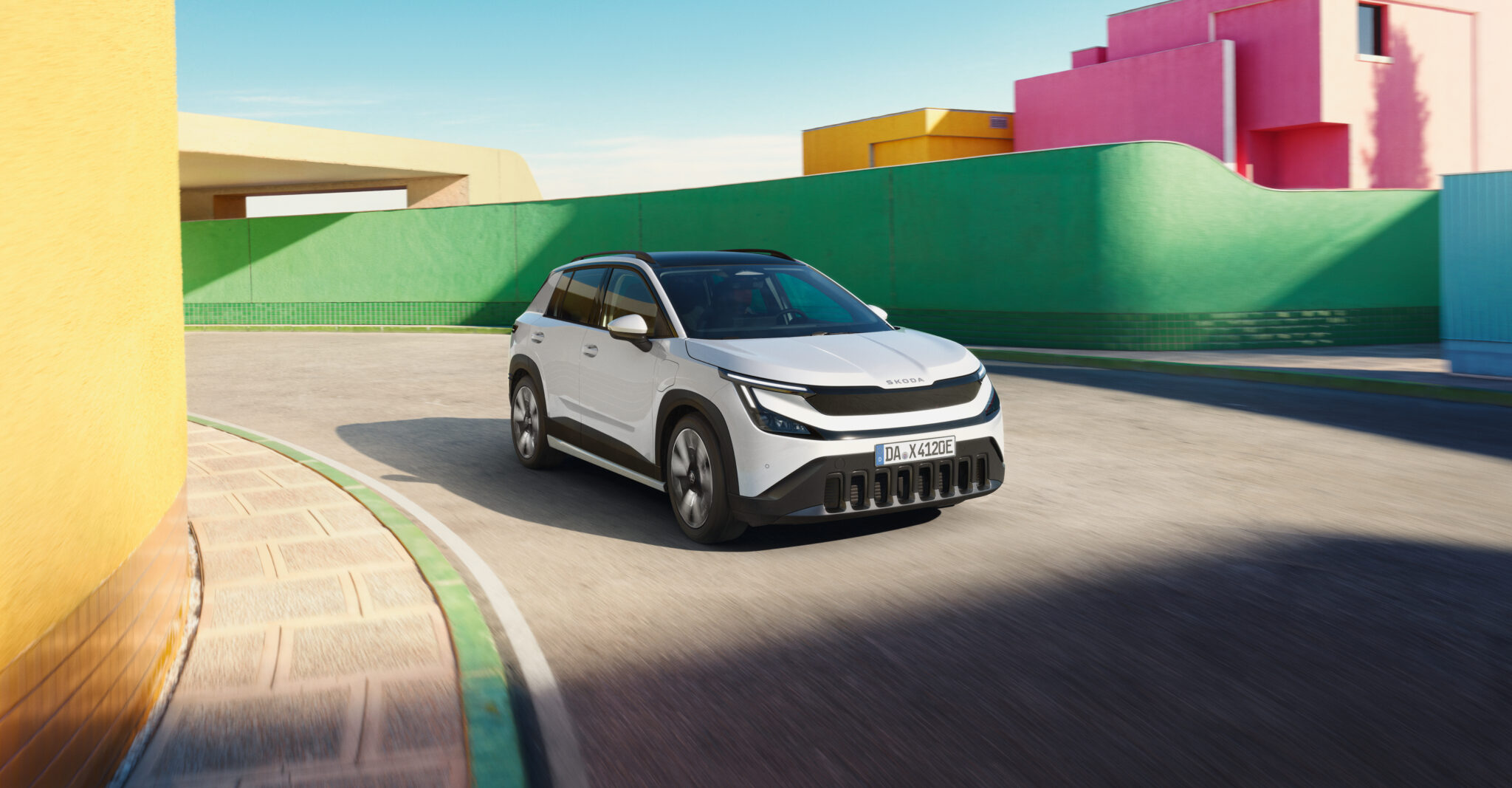 Ein weißer Skoda Epiq fährt in einer modern gestalteten Umgebung. Die Gebäude und Mauern im Hintergrund sind in kräftigen Farben wie Grün, Gelb und Pink gestaltet. Die Aufnahme vermittelt Bewegung und eine helle, sonnige Atmosphäre