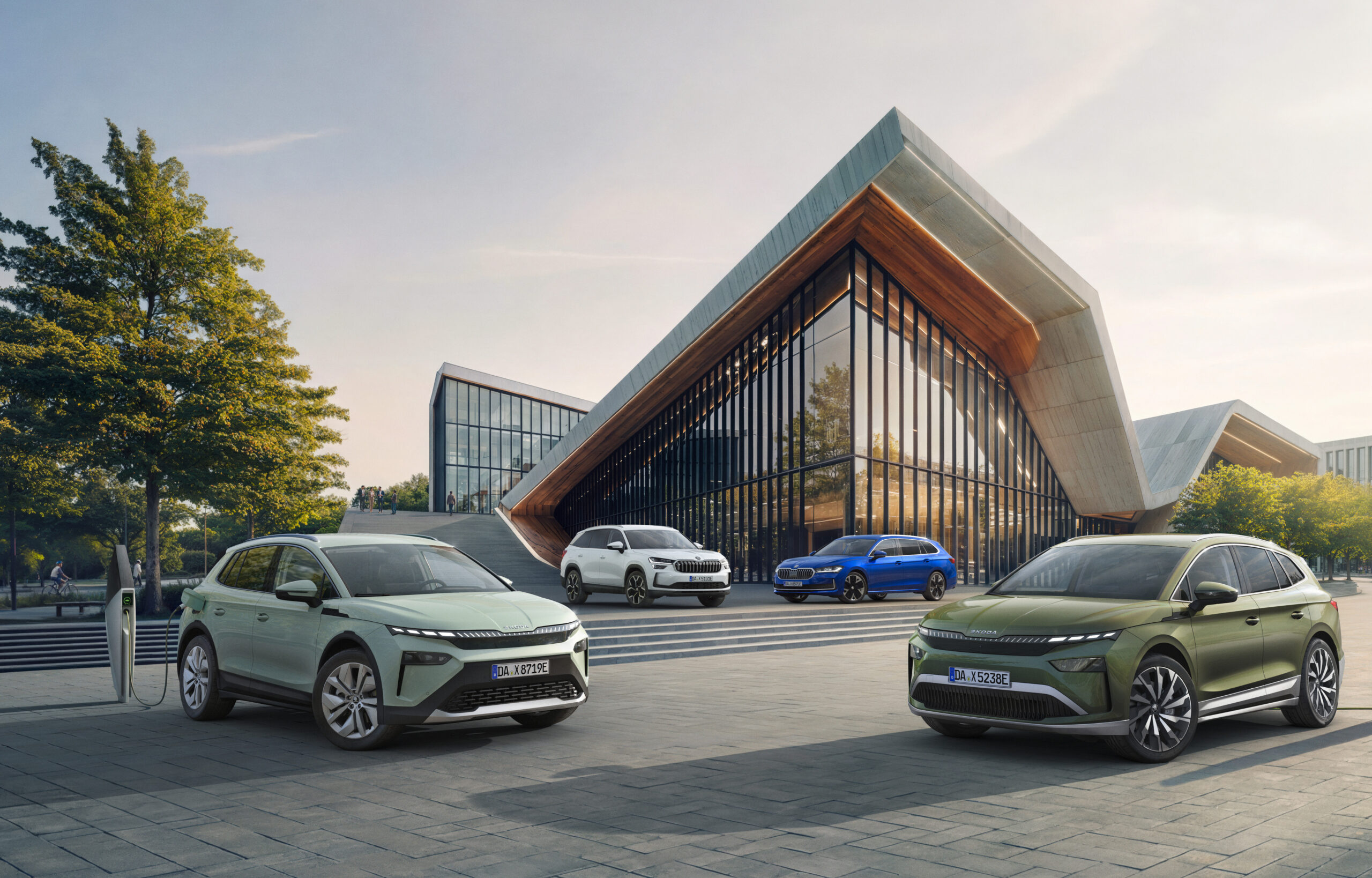 Auf dem Bild sind 4 Elektrofahrzeuge von Skoda in den Farben dunkelgrün, hellgrün, weiß und blau zu sehen. Die grünen Fahrzeuge sind an eine Ladesäule angeschlossen und stehen im Vordergrund. Die beiden anderen Fahrzeuge stehen im Hintergund auf einer Treppe vor einem großen luxuriösen Gebäuse mit einer Glasfront und einem beigen Dach. Links im Bild ist ein großer grüner Baum zu sehen.