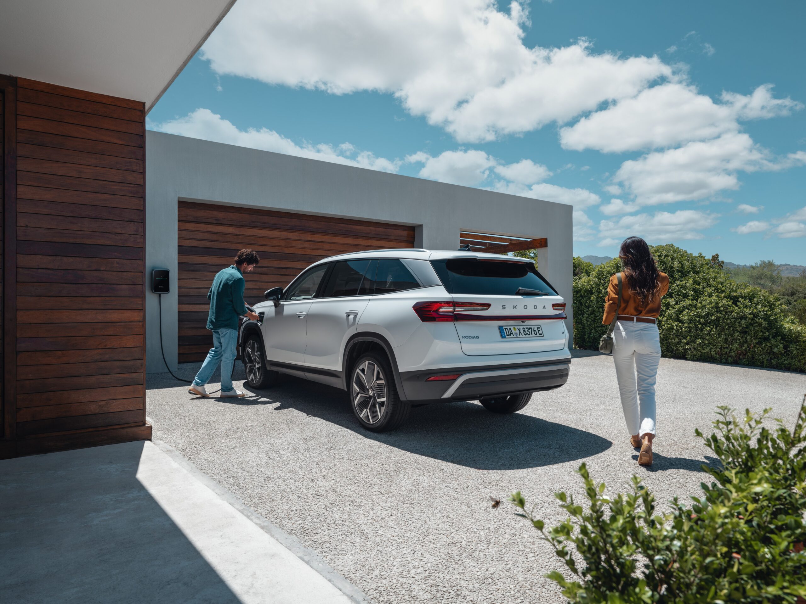 Auf den Bild ist der Skoda Kodiaq in der Farbe weiß und ein Mann, der das Elektrofahrzeug an die Ladestation anschließt zu sehen. Hinter dem Fahzeug läuft eine Dame. Das Fahrzueg steht vor einer Garange mit Holzoptik. Der Himmel ist blau und es sind ur wenige Wolken zu sehen. Das Bild wirkt hell und sommerlich.