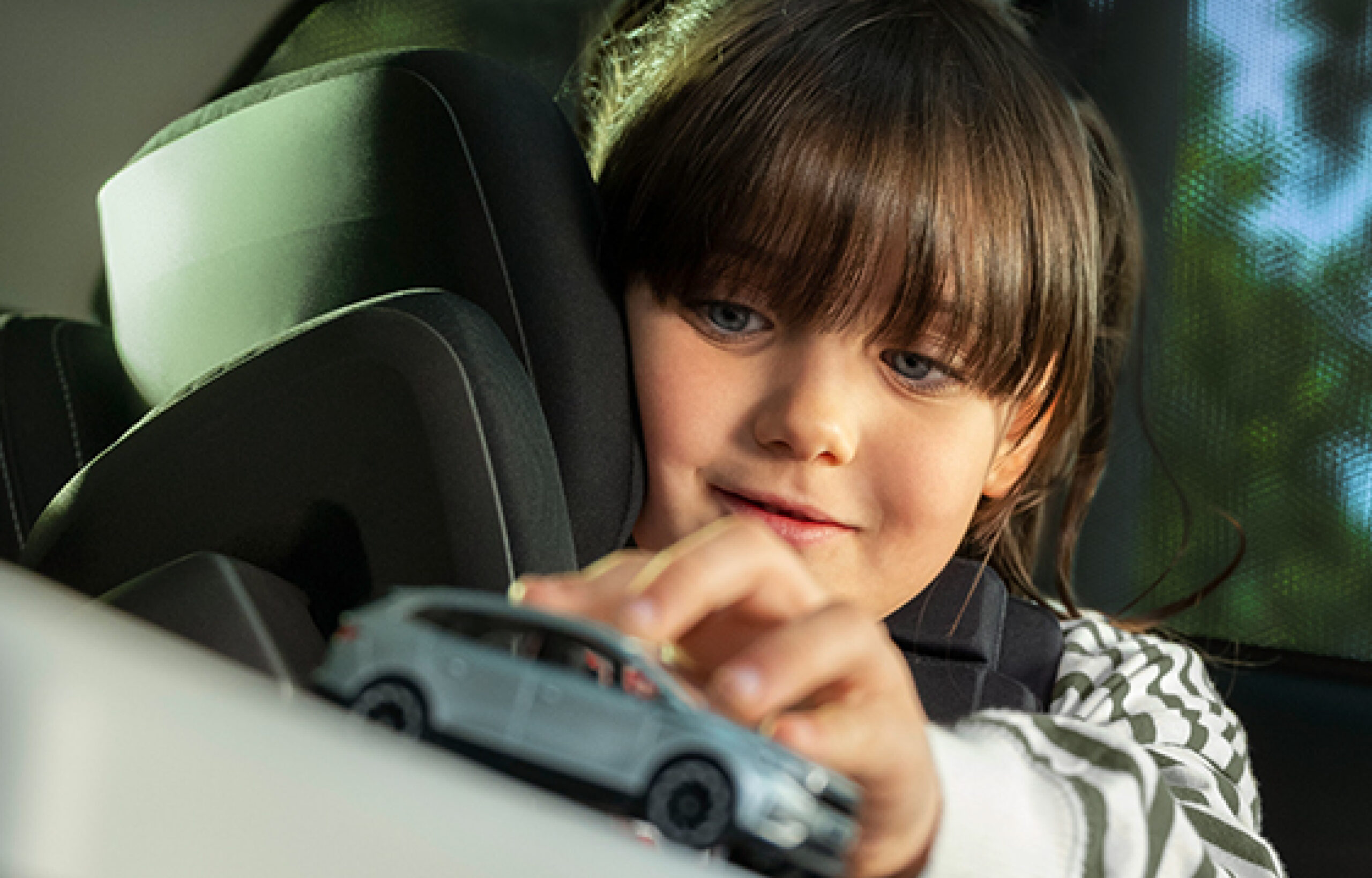 Ein kleines braunhaariges Mädchen sitzt in einem Kindersitz im Auto. Sie spielt mit einem Spielzeugauto.