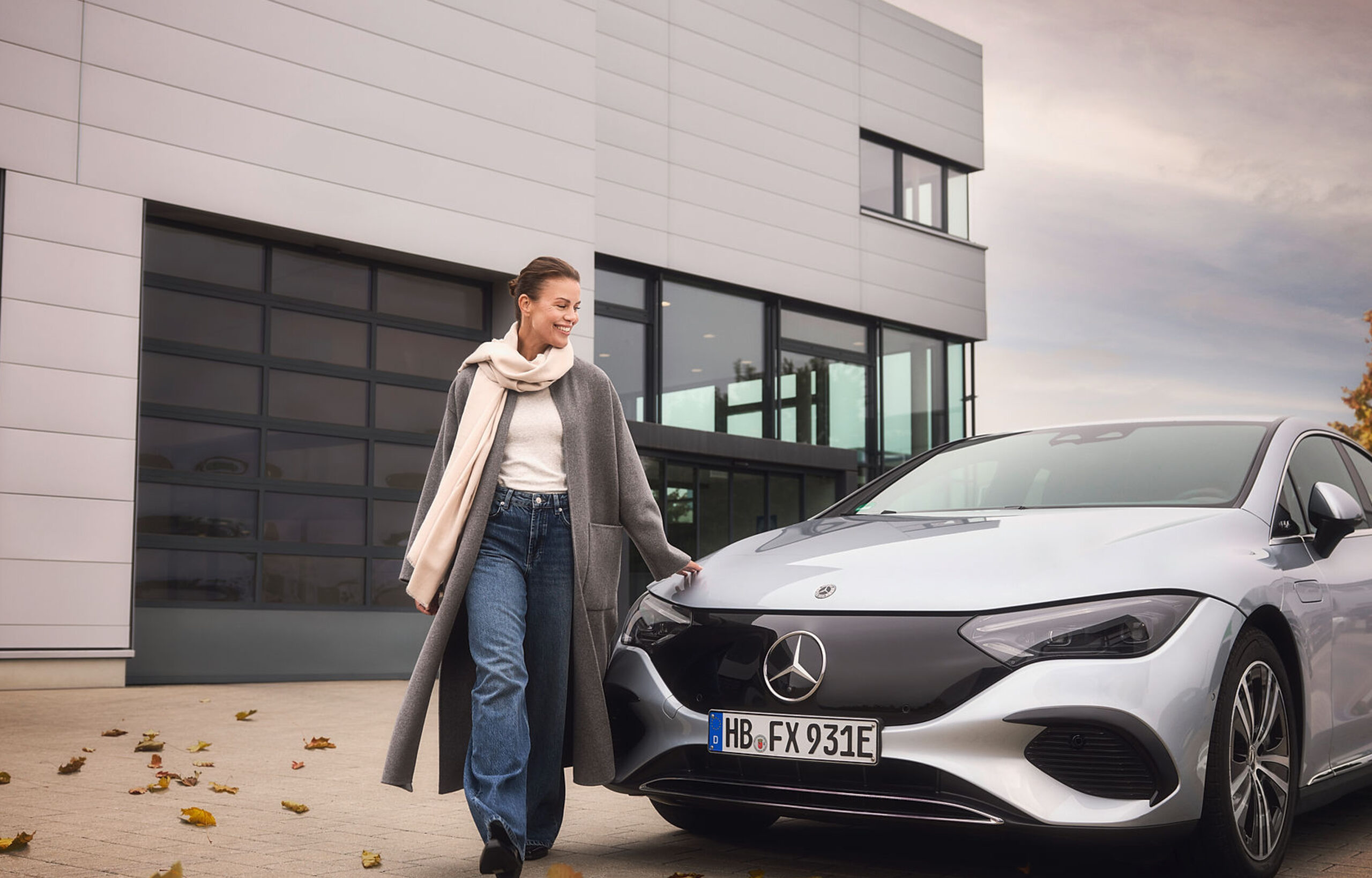 Eine Frau in langem Mantel und Schal läuft glücklich an ihrem Mercedes vorbei und legt die Hand auf. Herbstliche Stimmung.